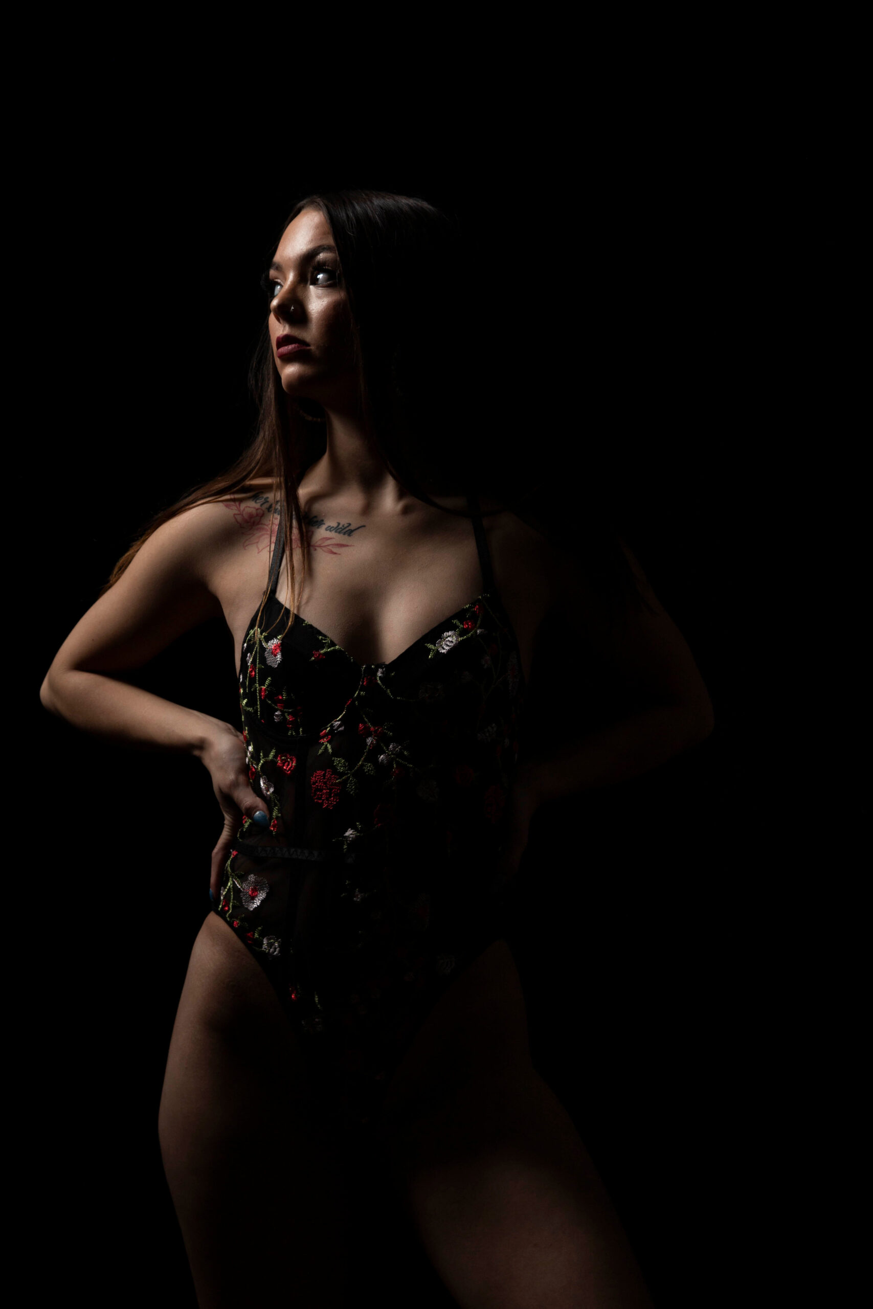 Shordy Kyy poses in a black floral bodysuit against a black studio background, with dramatic side lighting illuminating part of her face and figure.