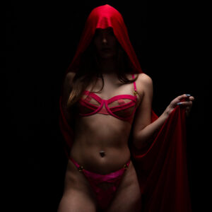 Shordy Kyy poses in pink lingerie beneath a flowing red hooded fabric against a completely black studio background.