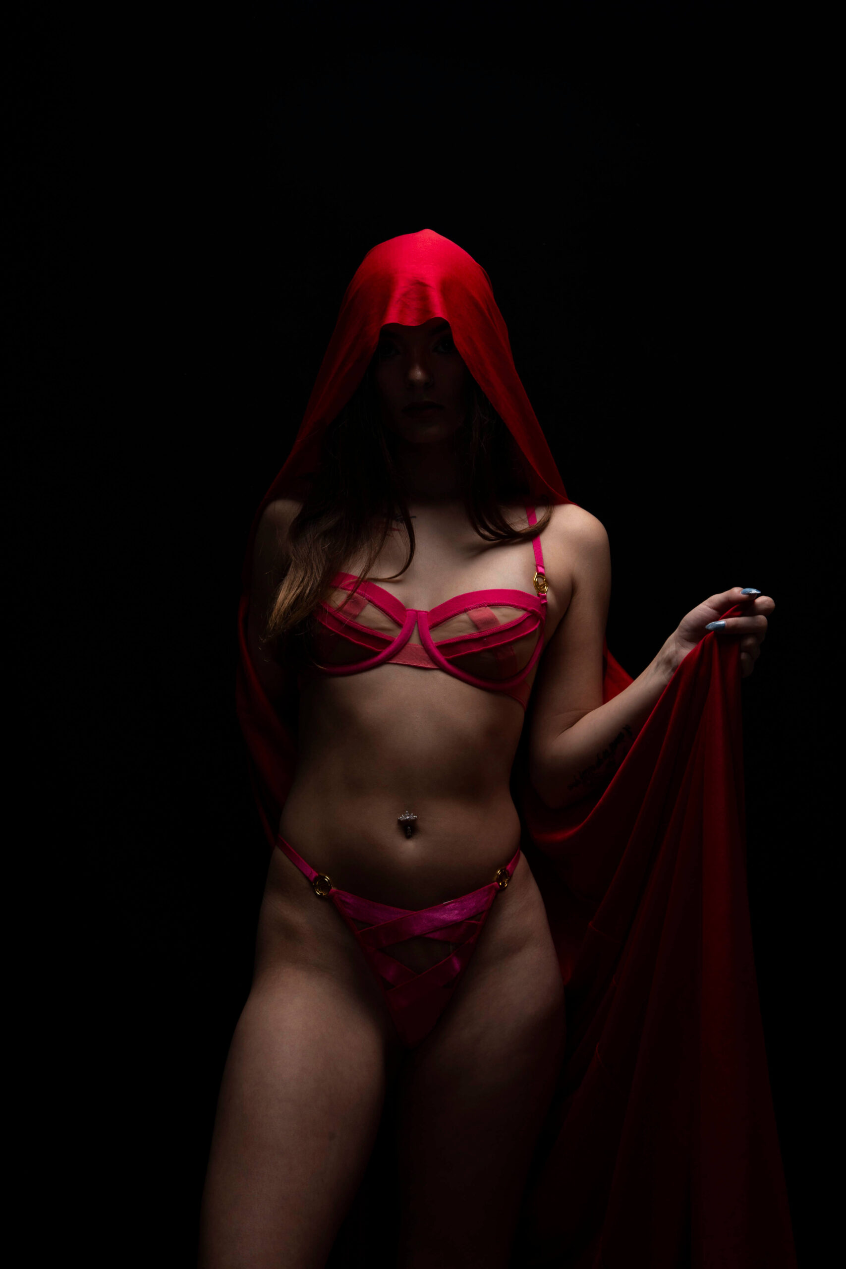 Shordy Kyy poses in pink lingerie beneath a flowing red hooded fabric against a completely black studio background.