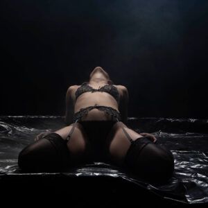 Shordy Kyy kneels in black lingerie and thigh-high stockings on a reflective black surface beneath dramatic overhead studio lighting.