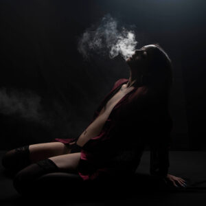 Shordy Kyy sits against a black studio background in dramatic low-key lighting while exhaling a cloud of smoke above her face.