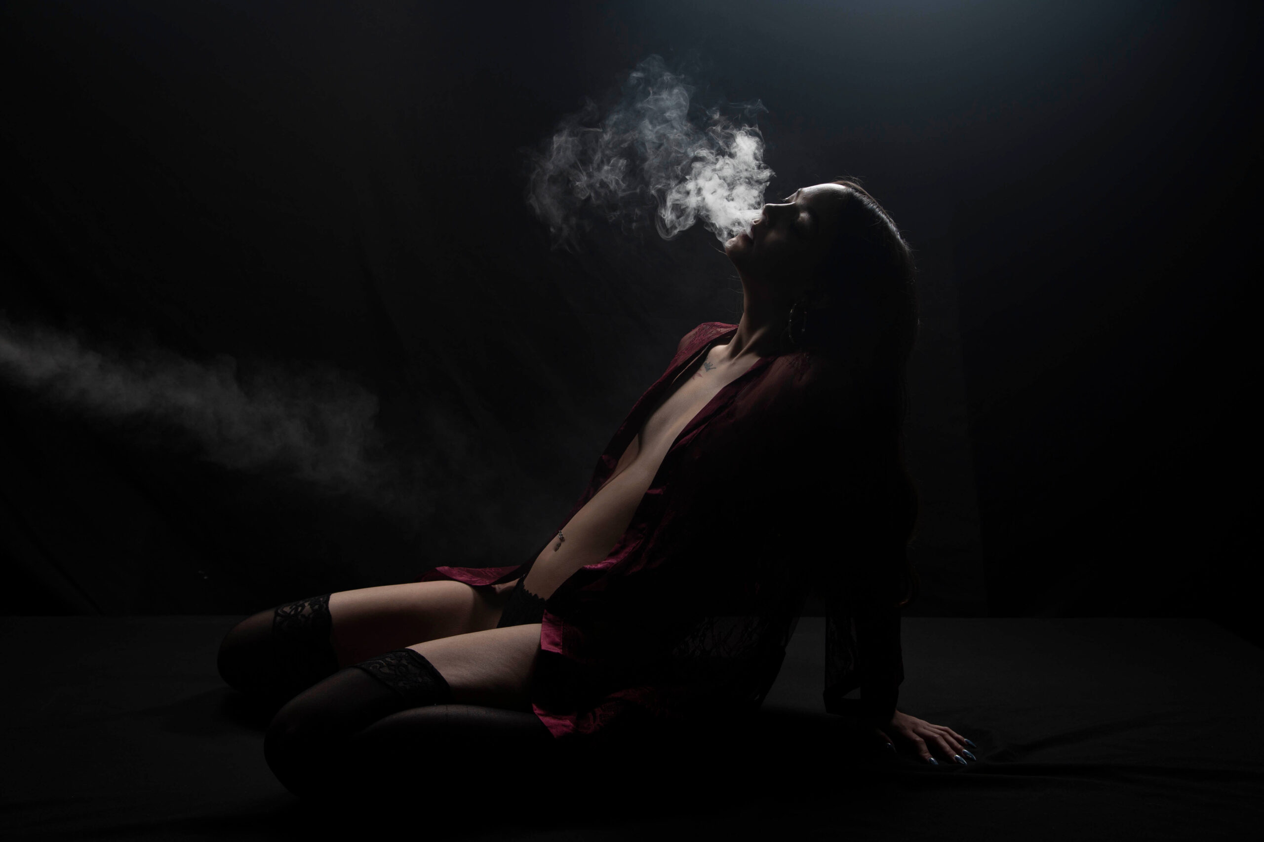 Shordy Kyy sits against a black studio background in dramatic low-key lighting while exhaling a cloud of smoke above her face.