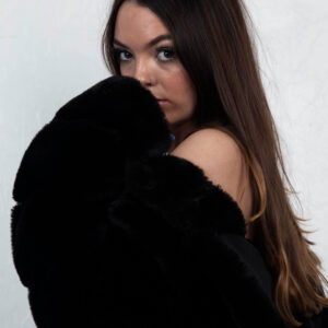 Shordy Kyy poses in a black faux-fur garment against a white studio background, looking directly toward the camera.