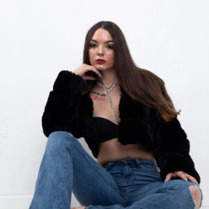 Shordy Kyy sits against a white studio wall wearing a black faux-fur jacket, black top, and distressed blue jeans.