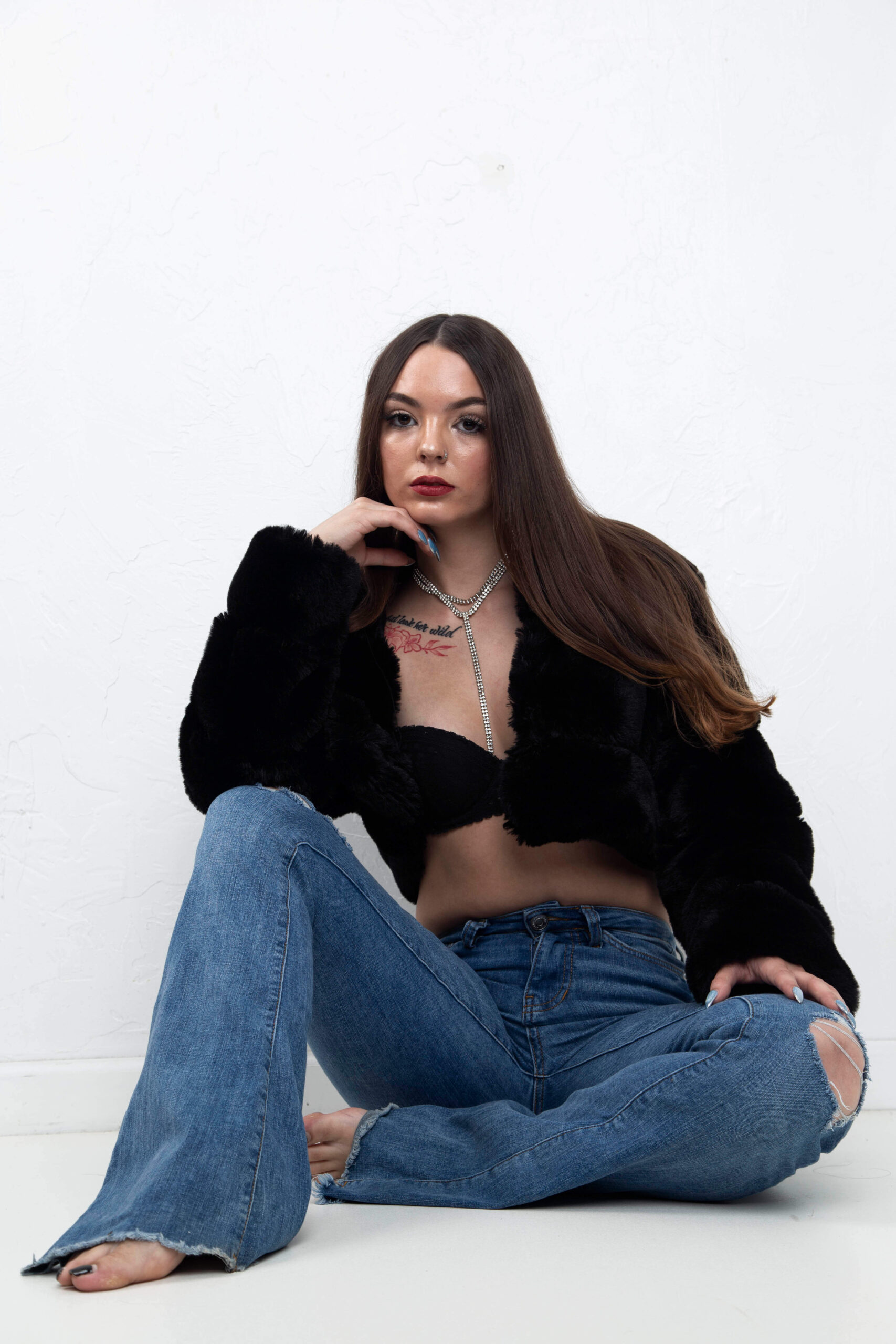 Shordy Kyy sits against a white studio wall wearing a black faux-fur jacket, black top, and distressed blue jeans.