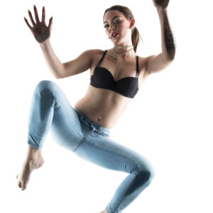 Shordy Kyy poses above a glass surface in a black bra and light blue jeans, with her hands raised against a bright white background.