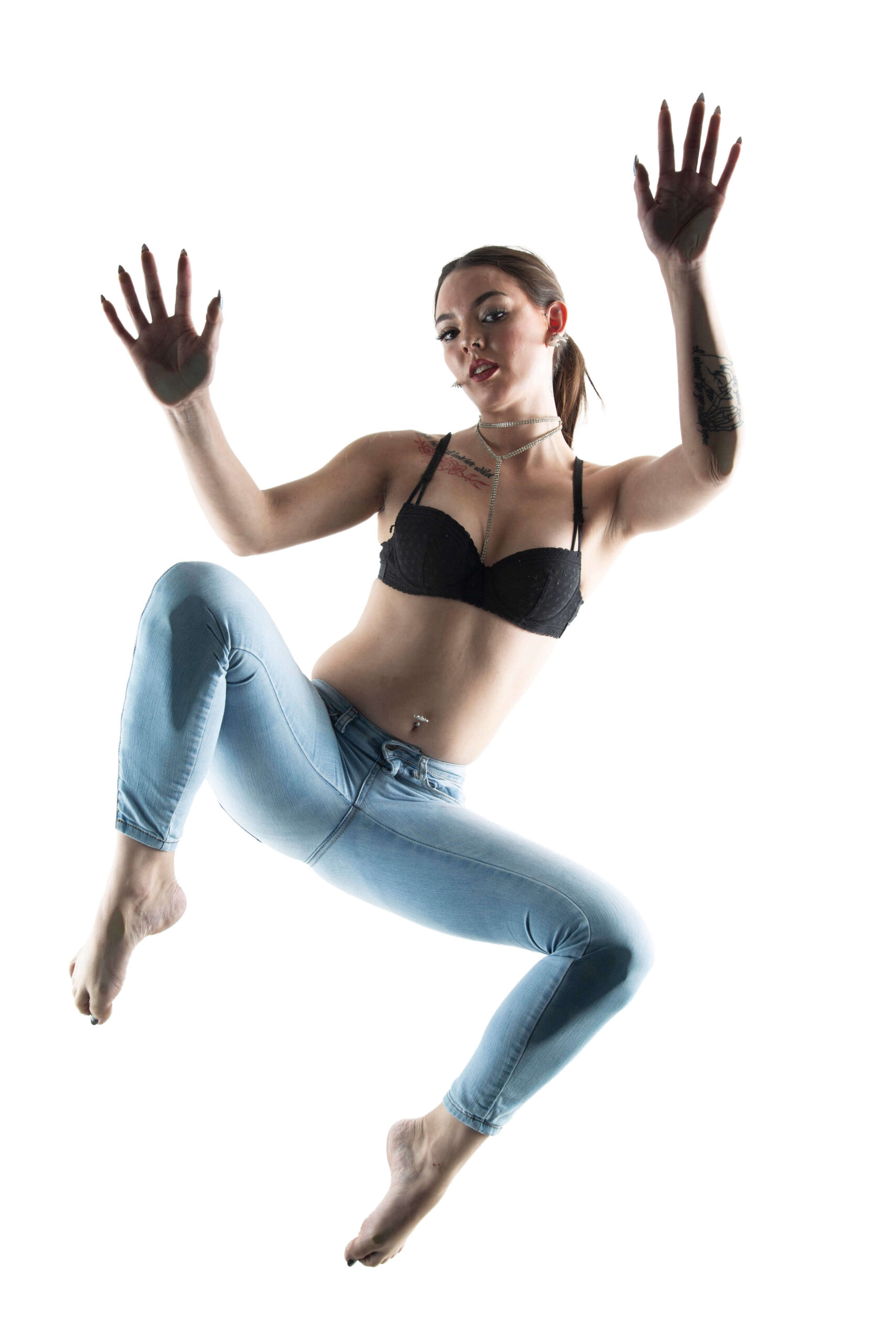 Shordy Kyy poses above a glass surface in a black bra and light blue jeans, with her hands raised against a bright white background.
