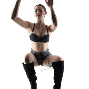 Shordy Kyy poses with both arms raised while wearing black lingerie, denim shorts, and tall black boots against a bright white studio background.