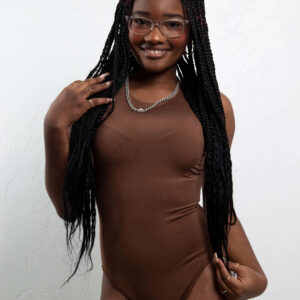 Fashion and glamour model Bri_Baddies14 smiling in clear-framed glasses and a fitted chocolate-brown bodysuit, holding her long black braids while posing against a textured white studio wall.