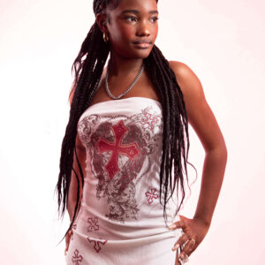 Fashion model Bri_Baddies14 wearing a fitted white strapless dress featuring decorative red crosses and intricate graphic patterns, posing with one hand on her hip against a softly illuminated pink and white studio background.