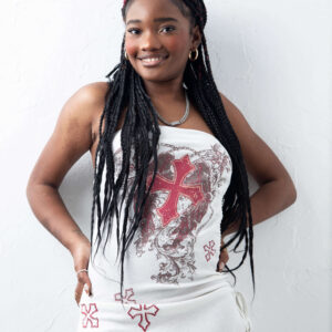 Fashion model Bri_Baddies14 smiling at the camera in a white strapless mini dress featuring red crosses and decorative graphics, with long black braids and gold hoop earrings against a textured white wall.