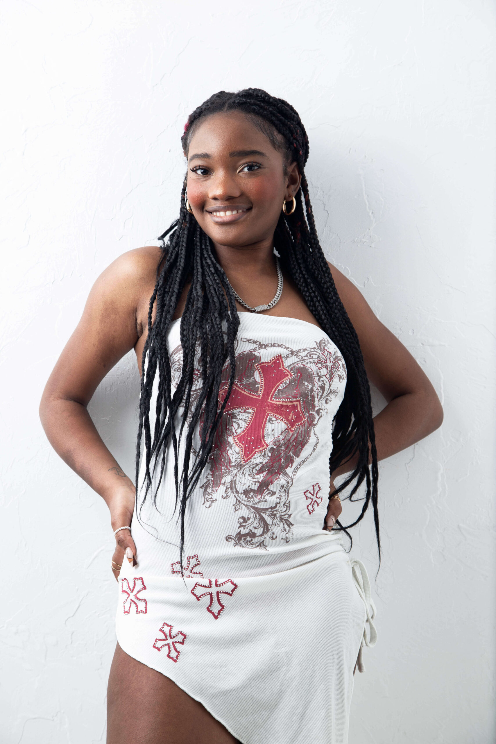 Fashion model Bri_Baddies14 smiling at the camera in a white strapless mini dress featuring red crosses and decorative graphics, with long black braids and gold hoop earrings against a textured white wall.