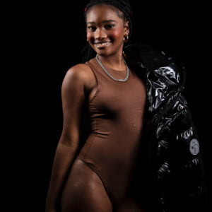 Glamour model Bri_Baddies14 smiling in a fitted brown bodysuit while holding a glossy black puffer jacket over her shoulder against a black studio background.