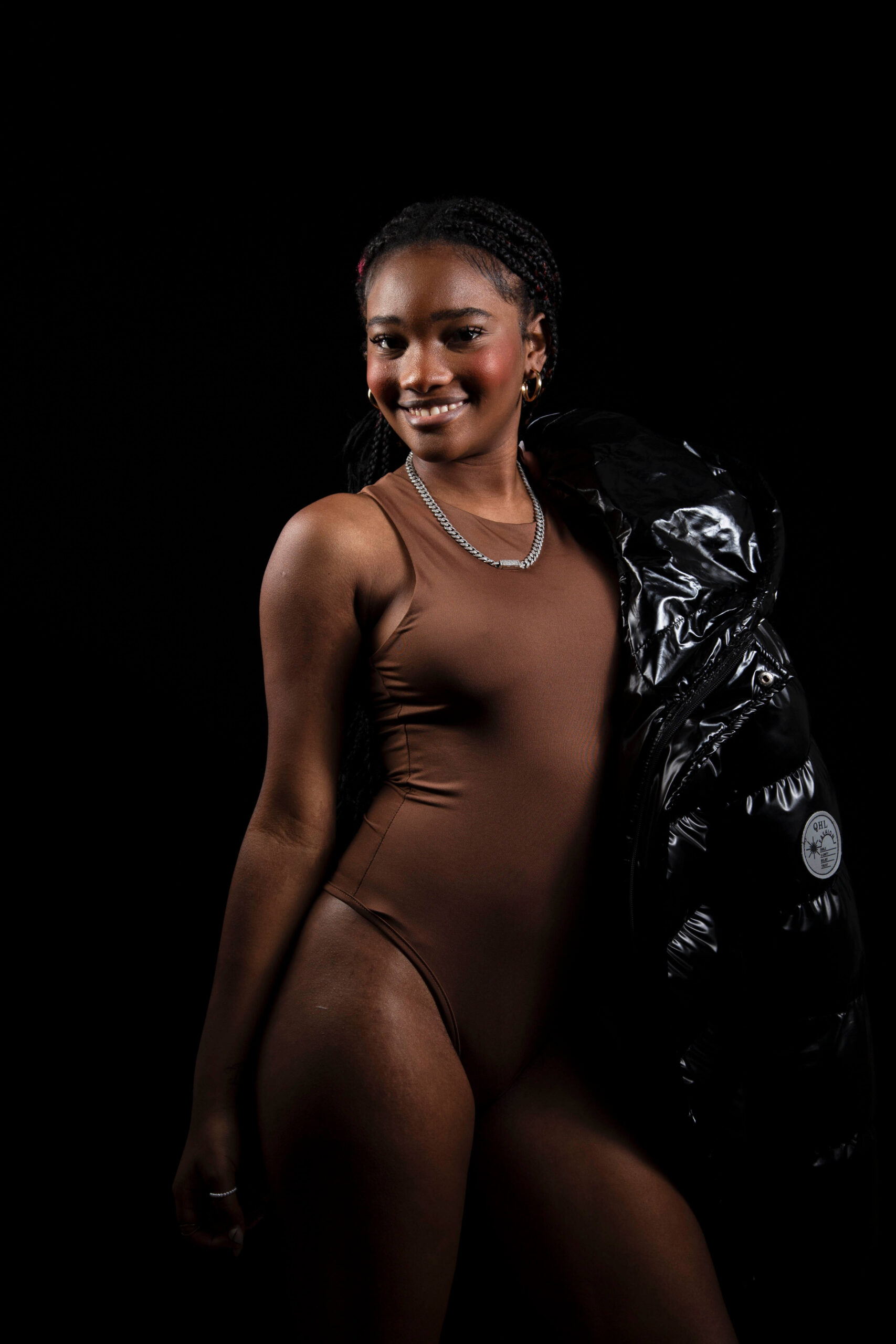 Glamour model Bri_Baddies14 smiling in a fitted brown bodysuit while holding a glossy black puffer jacket over her shoulder against a black studio background.