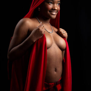 Artistic topless glamour portrait of model Bri_Baddies14 wearing a flowing red hooded cloak and red-and-black plaid bottoms, smiling against a dramatic black studio background.