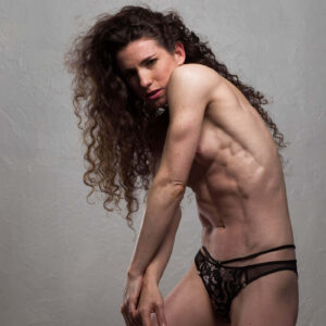 Keira Grant poses against a textured gray wall, wearing black lace bottoms with her long curly hair cascading over one shoulder.