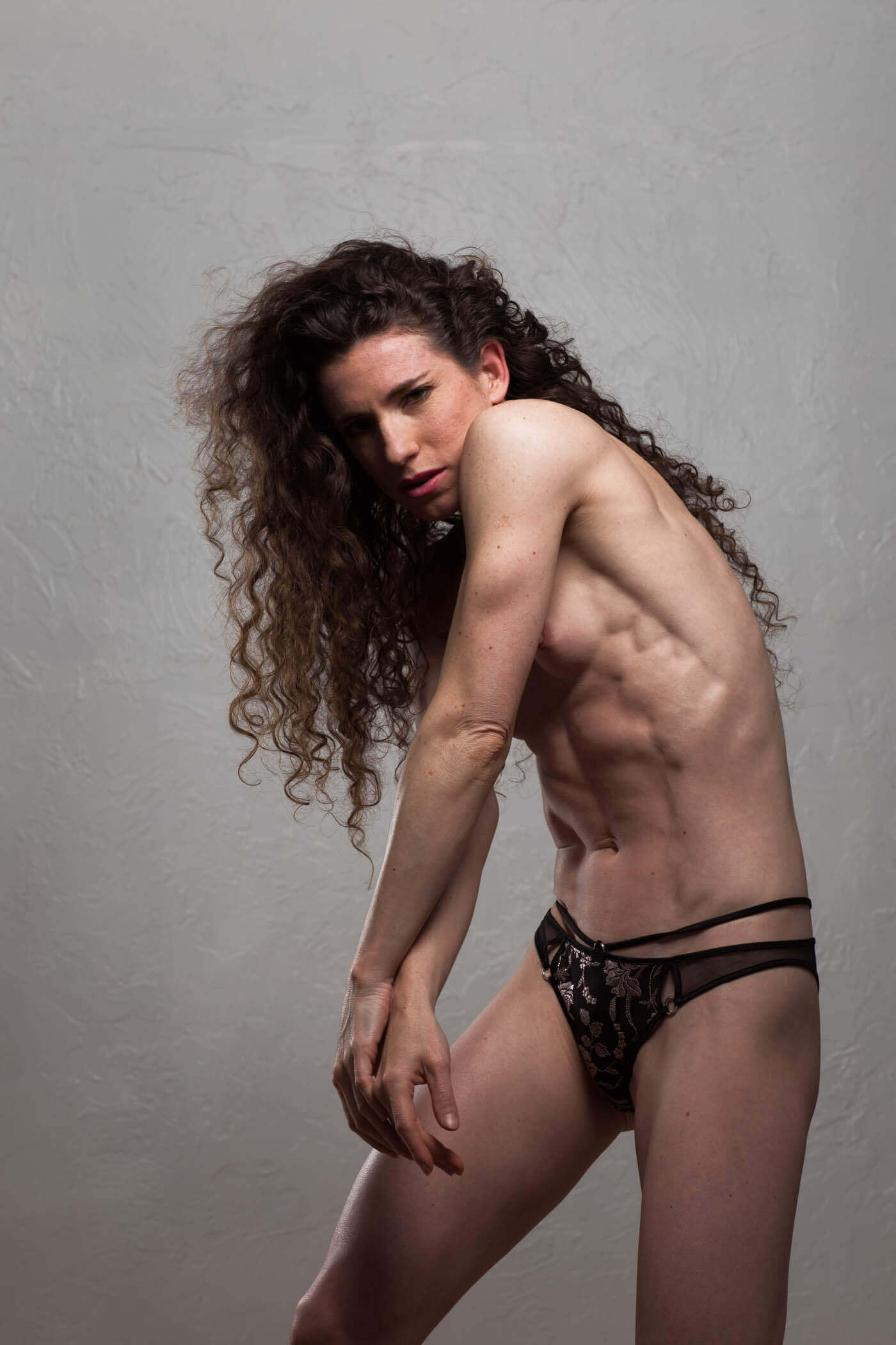Keira Grant poses against a textured gray wall, wearing black lace bottoms with her long curly hair cascading over one shoulder.