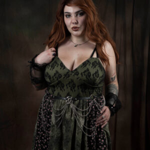 El The Belle poses against a dark brown studio backdrop wearing an olive green and black lace dress accented with a chain belt and black lace cuffs.