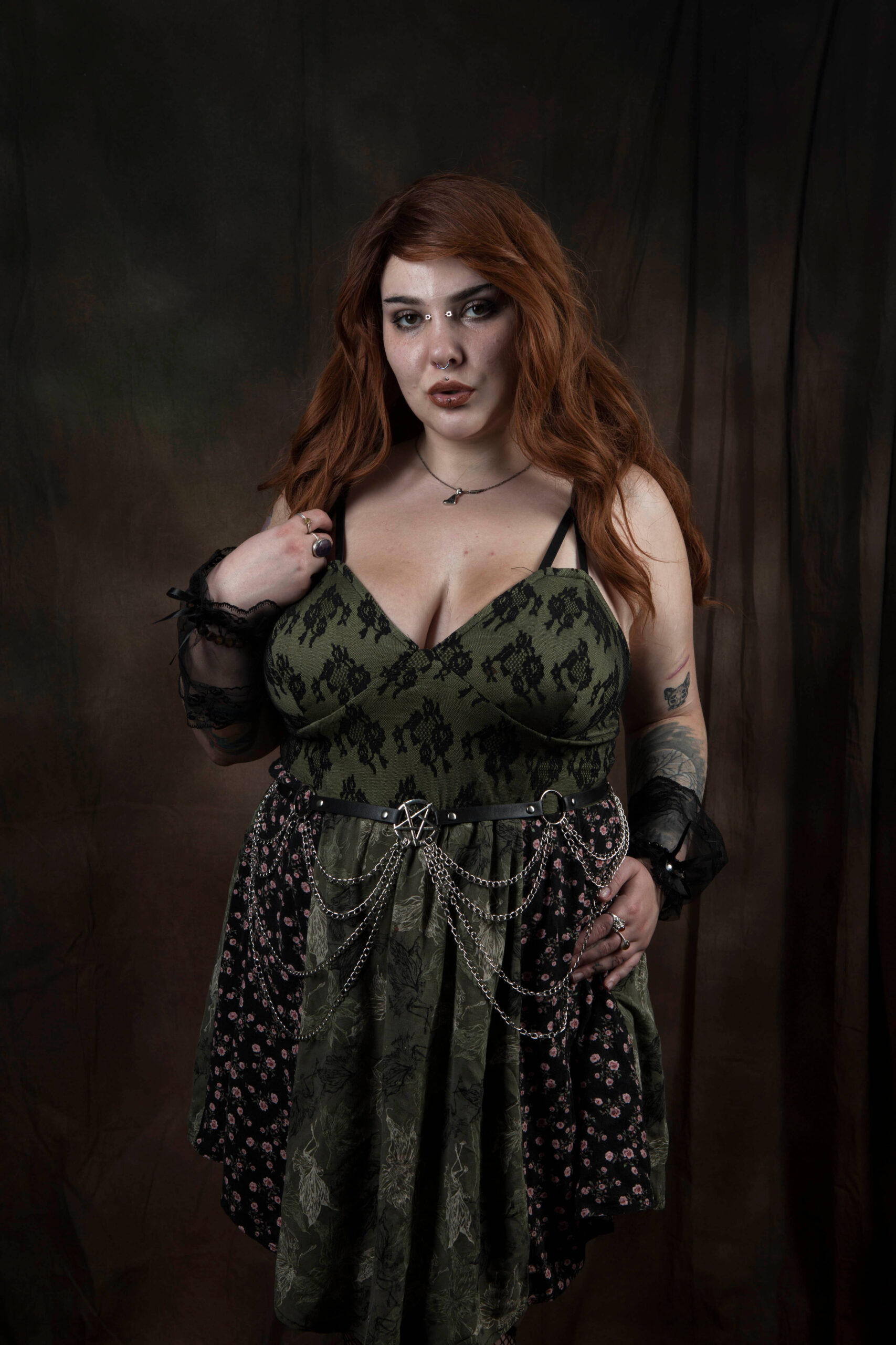 El The Belle poses against a dark brown studio backdrop wearing an olive green and black lace dress accented with a chain belt and black lace cuffs.