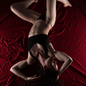 Keira Grant reclines in black lingerie against richly textured red fabric, forming an expressive pose with bent legs and flowing dark curls.