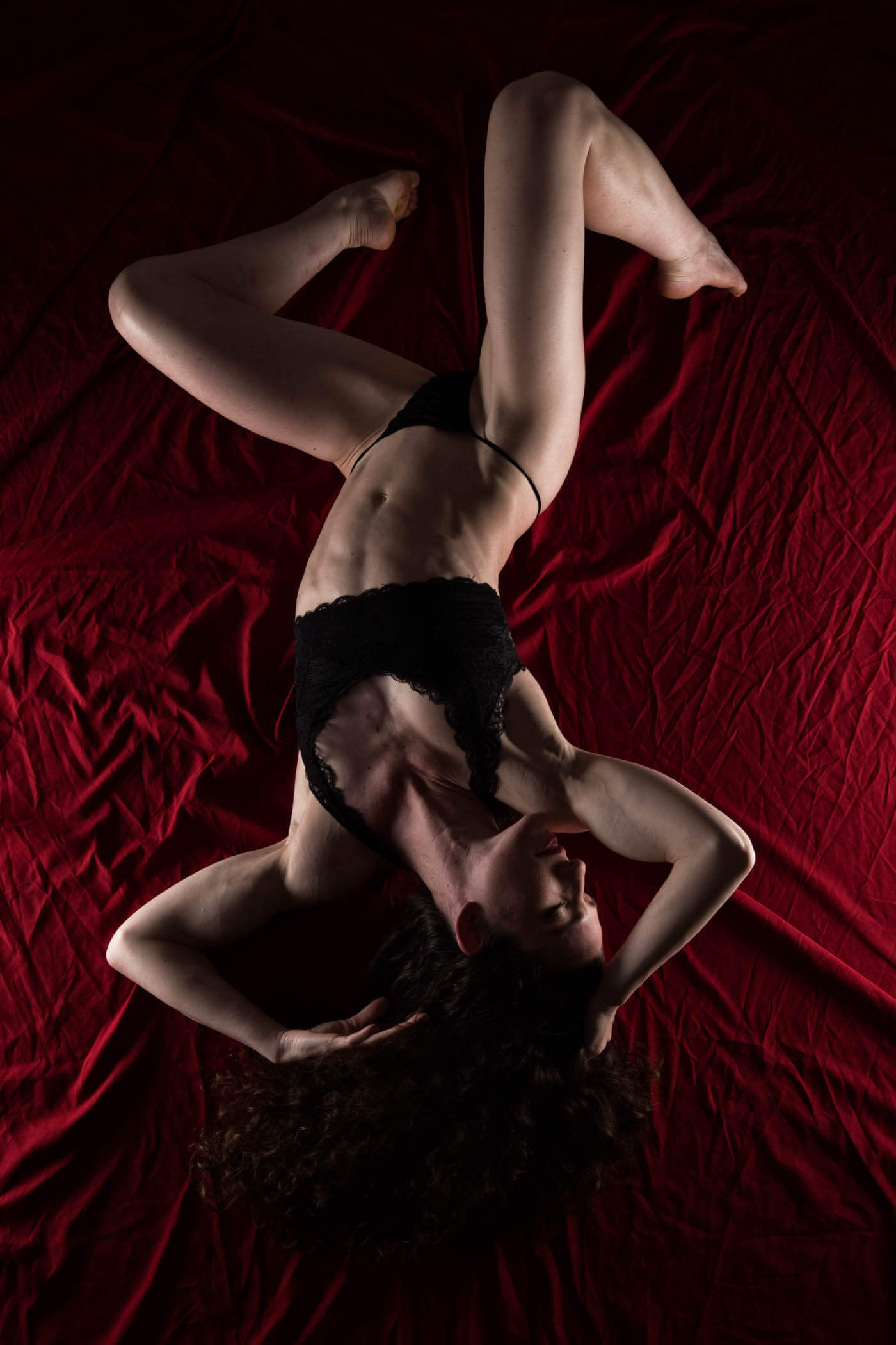 Keira Grant reclines in black lingerie against richly textured red fabric, forming an expressive pose with bent legs and flowing dark curls.