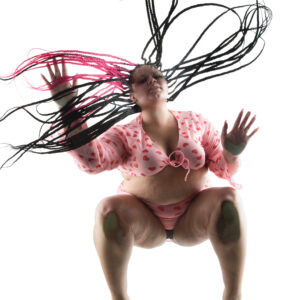 Kylee the Model poses above a glass platform in a pink heart-patterned outfit, with long black and pink braids spreading dramatically across the frame.
