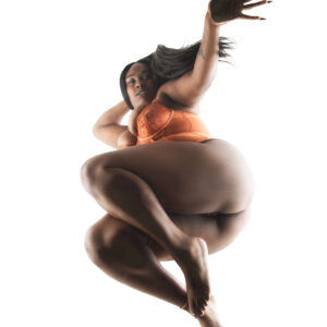 2Litt Modeling poses in orange lingerie with her legs crossed and one arm reaching toward the camera against a bright white background.