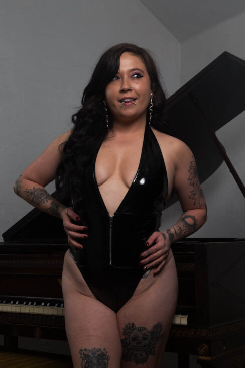 Oklahoma model Ganja Queen poses in a plunging black vinyl bodysuit in front of a dark antique piano, displaying her tattoos and facial piercings.