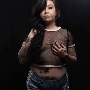 Ganja Queen poses against a black studio background wearing a sheer black mesh top and distressed denim shorts, with long dark hair, facial piercings and extensive tattoos visible.