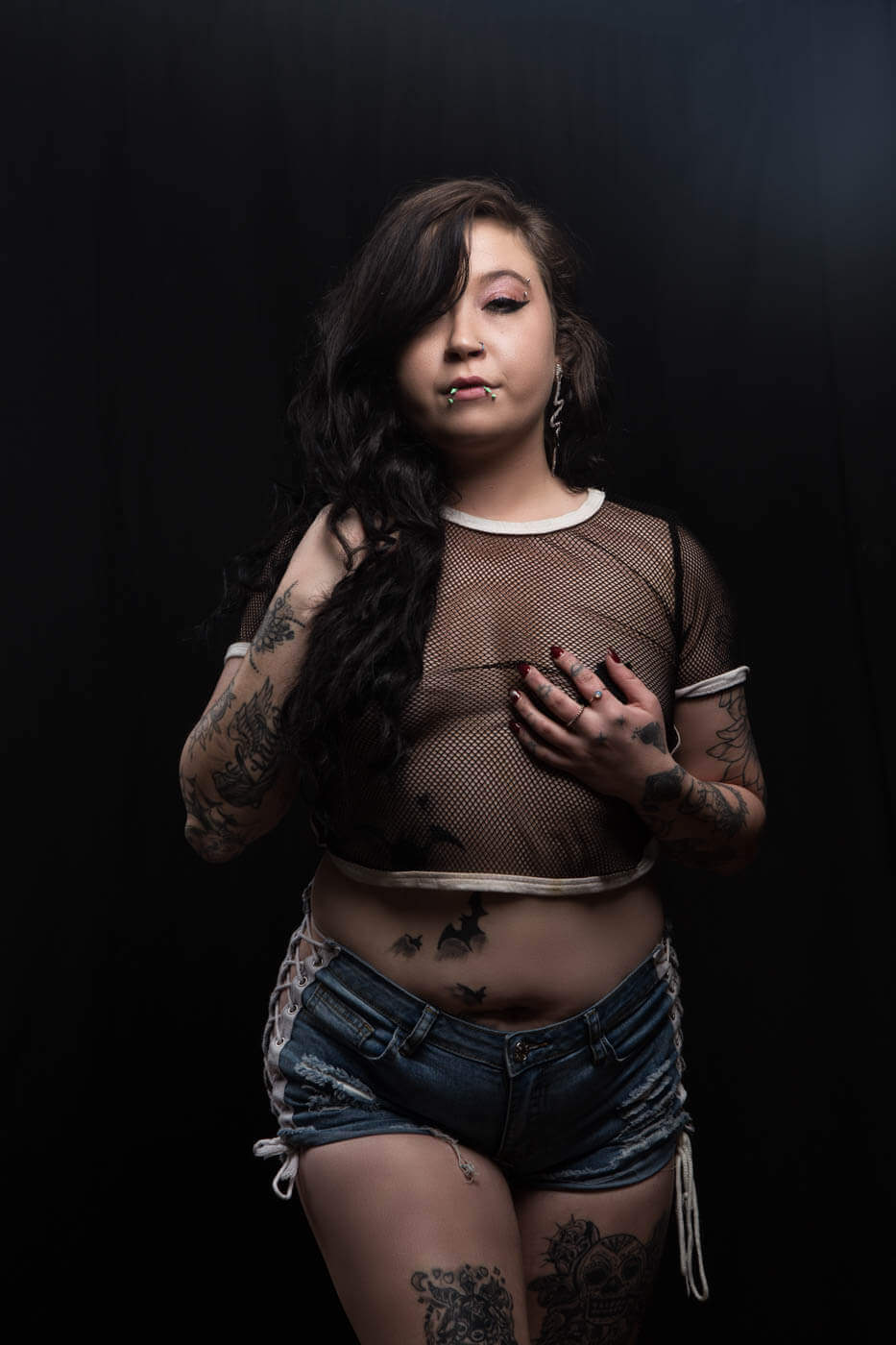 Ganja Queen poses against a black studio background wearing a sheer black mesh top and distressed denim shorts, with long dark hair, facial piercings and extensive tattoos visible.