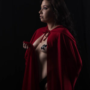 Ganja Queen poses in profile against a black studio background, draped in a vivid red hooded cape with long dark hair, dramatic makeup and facial piercings.