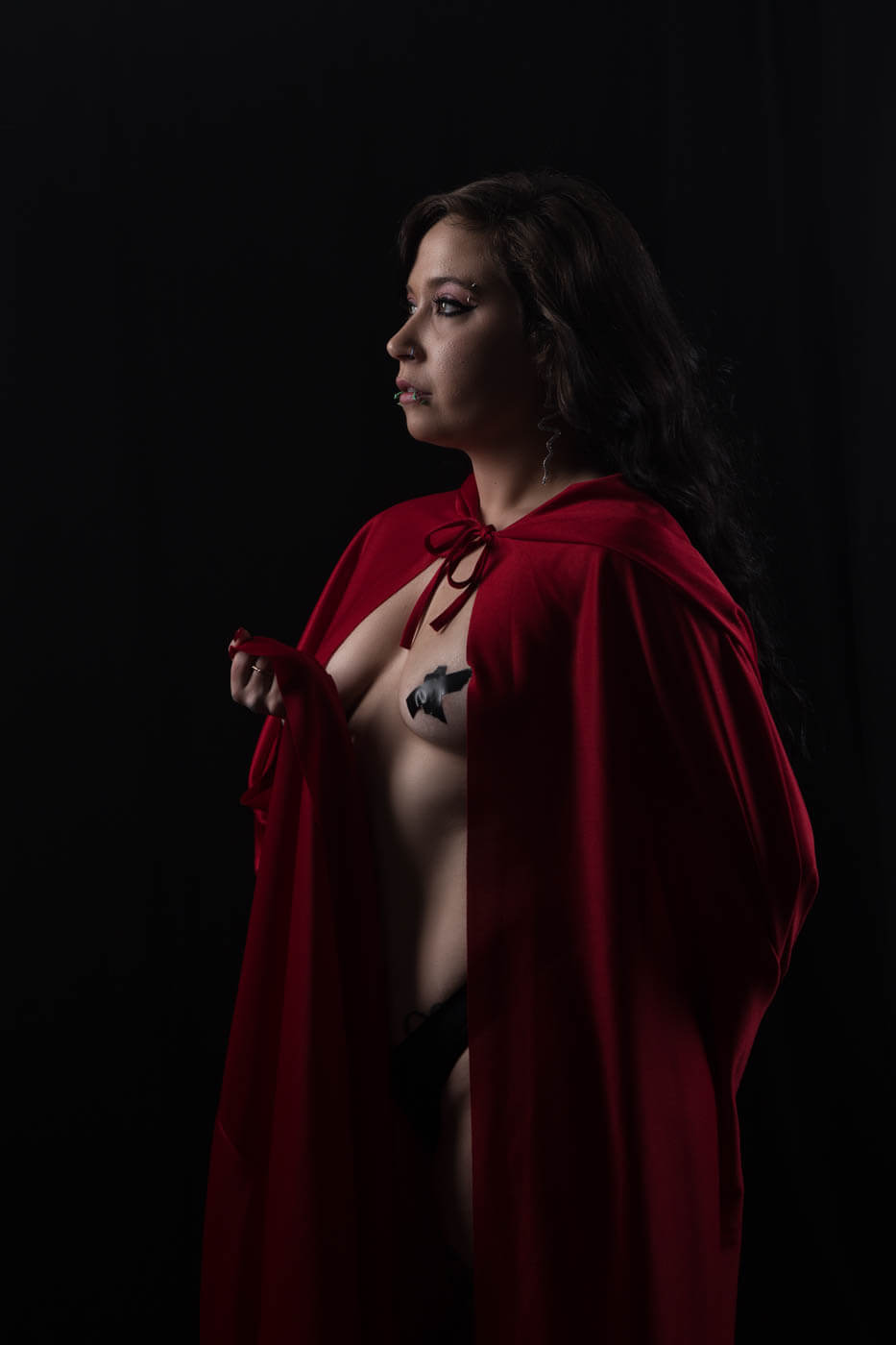 Ganja Queen poses in profile against a black studio background, draped in a vivid red hooded cape with long dark hair, dramatic makeup and facial piercings.