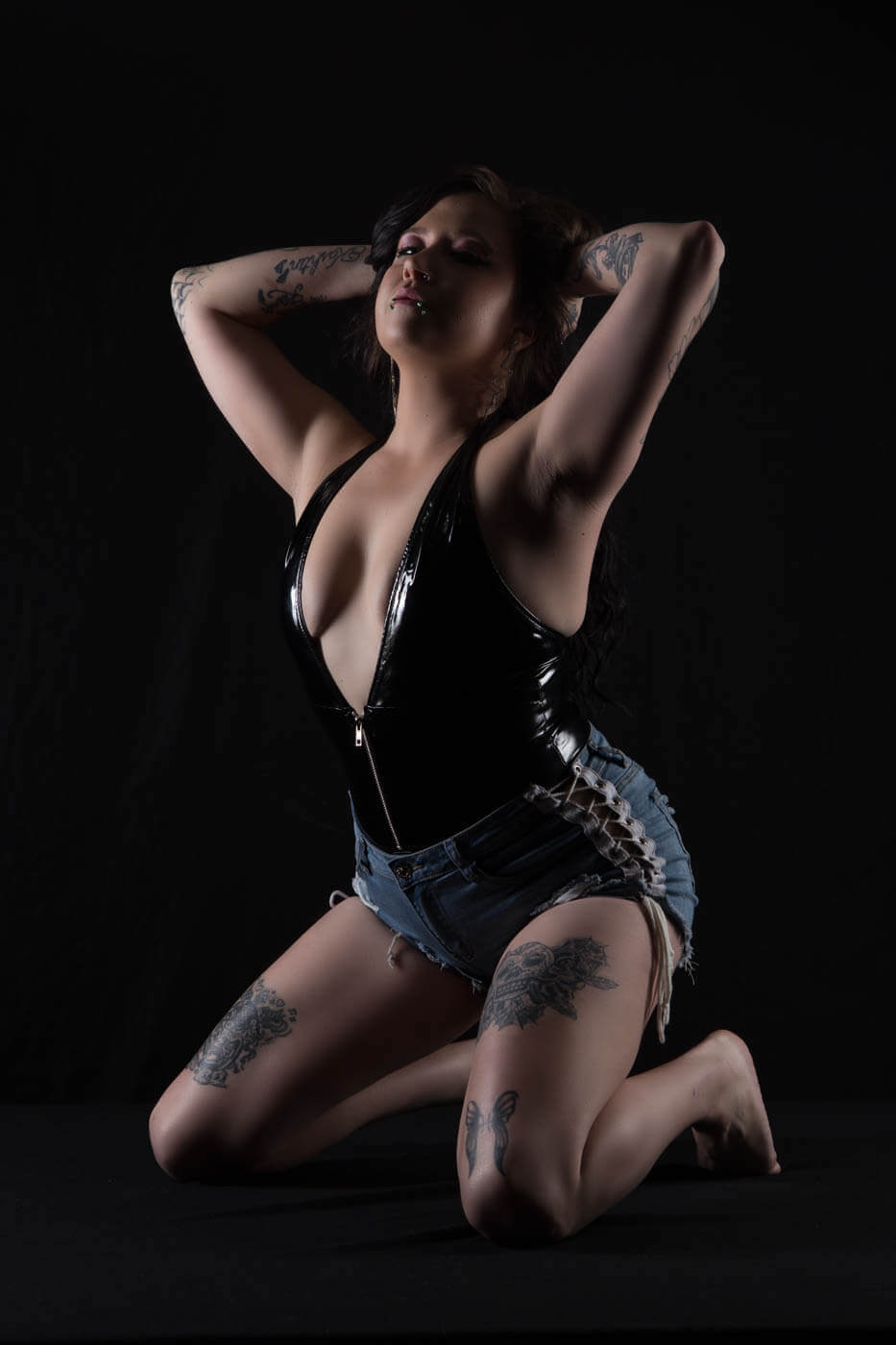 Ganja Queen kneels against a black studio background wearing a glossy black bodysuit and distressed denim shorts, with her arms raised behind her head and tattoos visible across her arms and legs.
