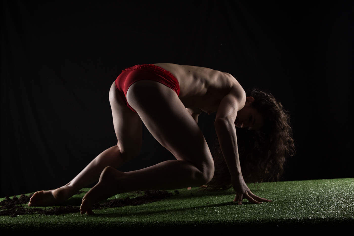 Keira Grant poses low to the ground in red bottoms on artificial grass, dramatically lit against a black background with scattered soil around her.