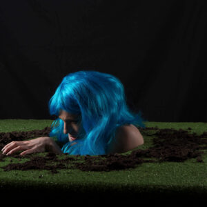 Keira Grant emerges through a dirt-covered opening in a patch of artificial grass, wearing a vivid blue wig against a dark black background.