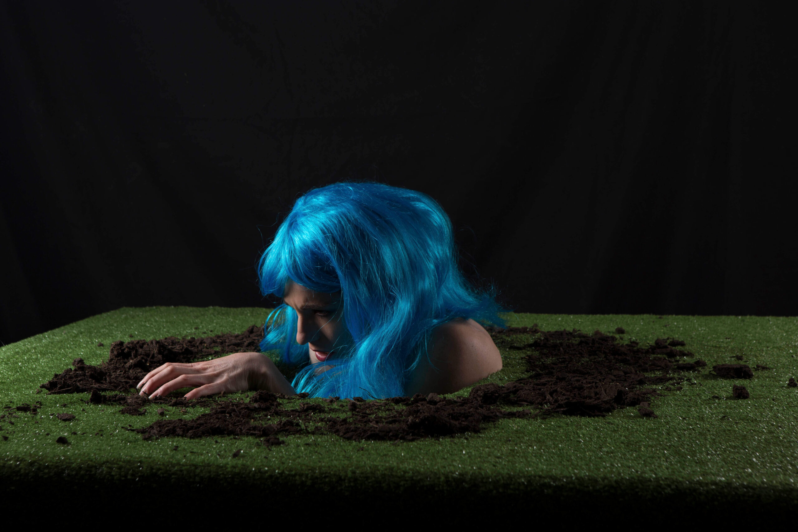 Keira Grant emerges through a dirt-covered opening in a patch of artificial grass, wearing a vivid blue wig against a dark black background.