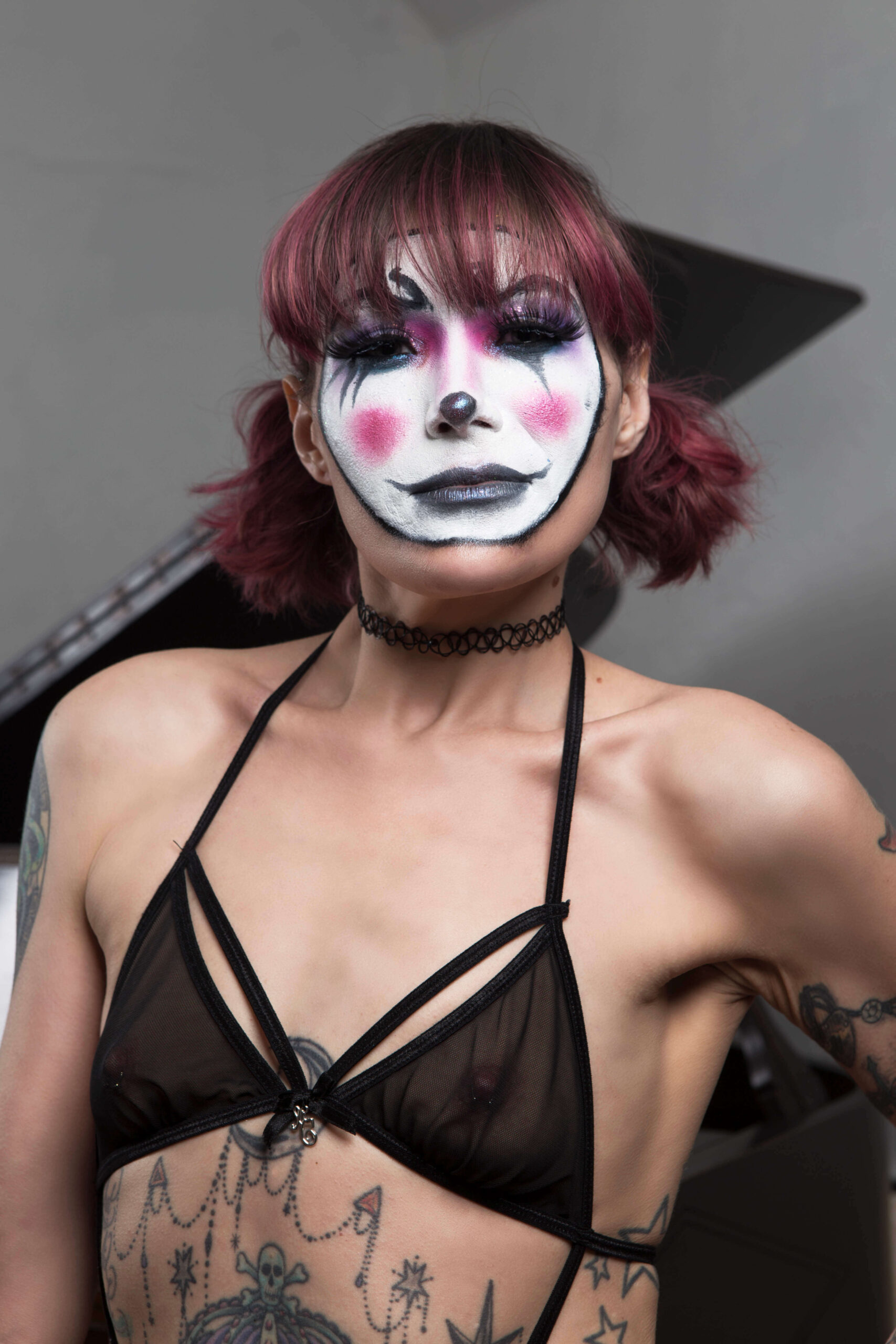 Kody Kathleen poses in a close-up portrait wearing dramatic black, white, and pink clown-inspired makeup, sheer black lingerie, a black choker, and her hair styled in pigtails.