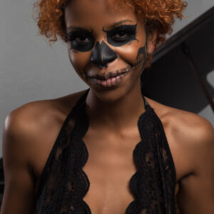 Close-up portrait of Saturn wearing dramatic black skull-inspired face paint, a black lace halter bodysuit, and short copper-colored curls.