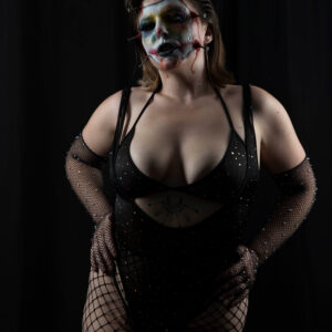 Alternative glamour model Birdie poses against a black studio backdrop wearing dramatic stitched horror makeup, a sparkling black bodysuit, fishnet sleeves, and fishnet stockings.
