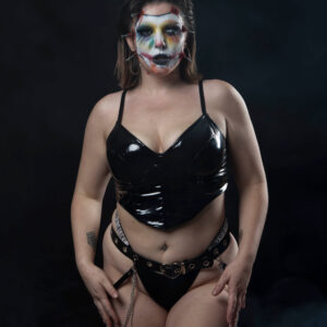 Alternative glamour model Birdie poses against a dark studio backdrop wearing elaborate horror-inspired face paint, simulated facial stitches, a glossy black vinyl corset top, black bottoms, and a decorative chain belt.