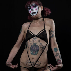 Kody Kathleen poses against a black backdrop wearing dramatic white, black, and pink clown-inspired makeup, sheer black lingerie, fishnet stockings, and a black choker.
