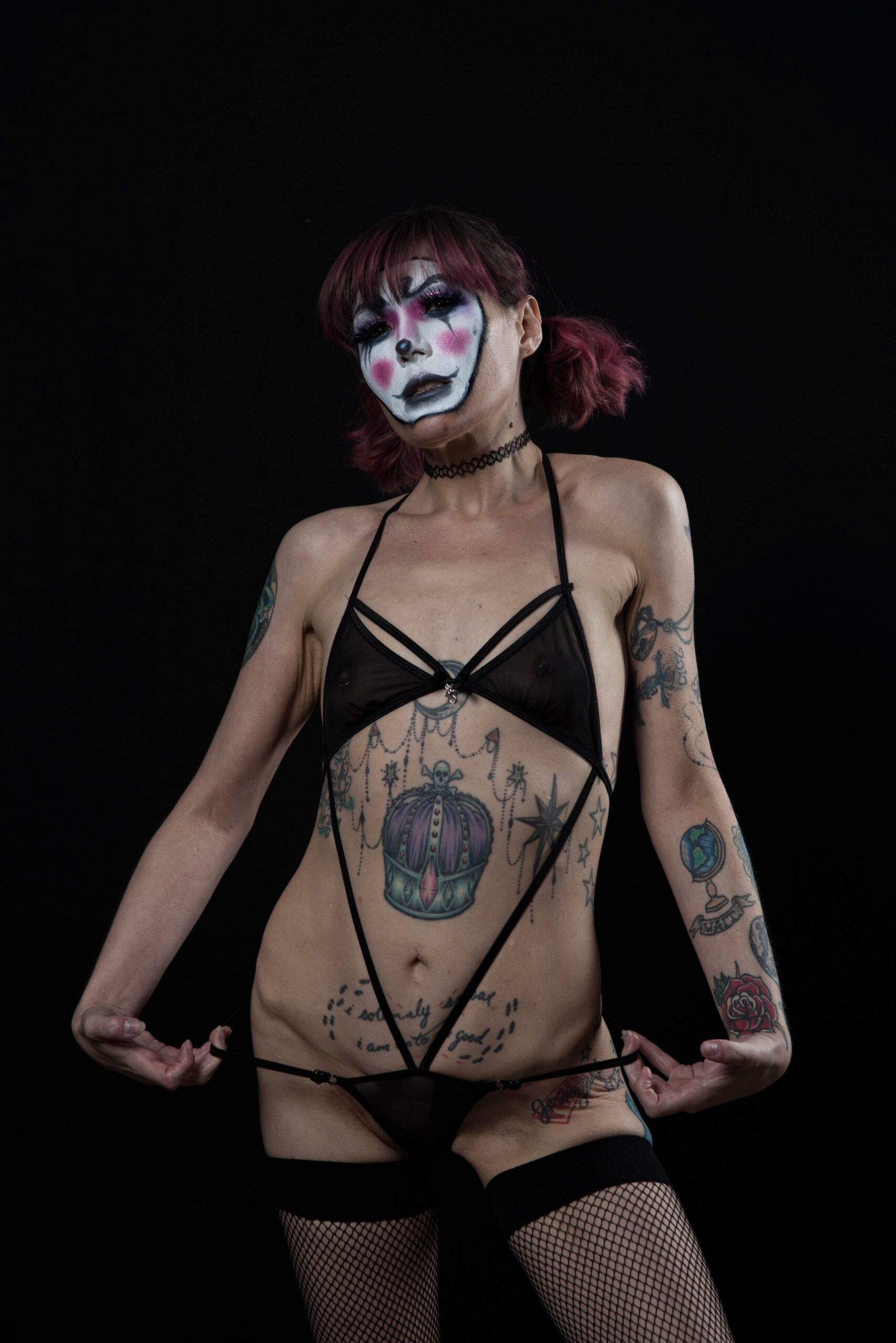 Kody Kathleen poses against a black backdrop wearing dramatic white, black, and pink clown-inspired makeup, sheer black lingerie, fishnet stockings, and a black choker.