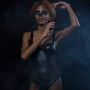 Saturn poses in a smoky dark studio wearing a black bodysuit and fishnet tights, with skull-inspired face paint and her arms raised in an expressive pose.