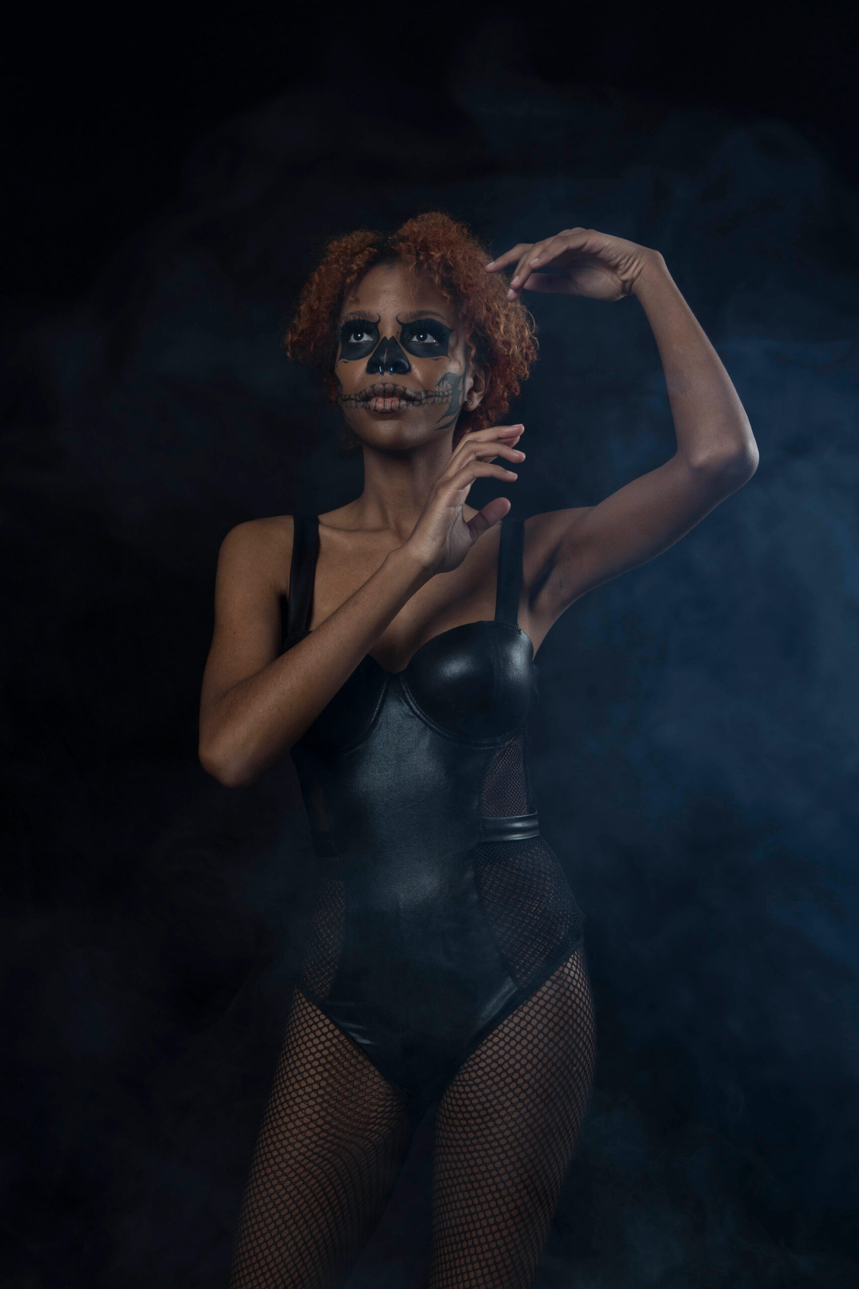 Saturn poses in a smoky dark studio wearing a black bodysuit and fishnet tights, with skull-inspired face paint and her arms raised in an expressive pose.