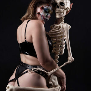 Alternative glamour model Birdie embraces a life-sized skeleton against a black studio backdrop. Wearing theatrical stitched horror makeup, a glossy black vinyl top, and strappy black bottoms with decorative chains, she poses in an intimate, gothic-inspired composition.