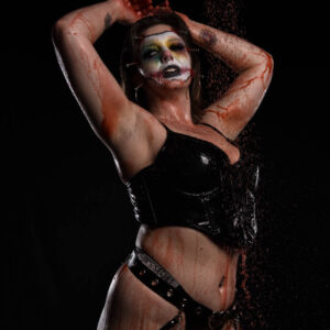 Alternative glamour model Birdie poses beneath a shower of crimson-colored liquid against a black studio backdrop. Wearing elaborate stitched horror makeup, a glossy black vinyl top, and strappy black bottoms with metallic chains, she raises both arms above her head in a dramatic gothic portrait.