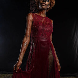 Saturn poses against a black background in a deep red lace dress with skull-inspired face paint as red liquid falls from above across her hair, shoulder, and clothing.
