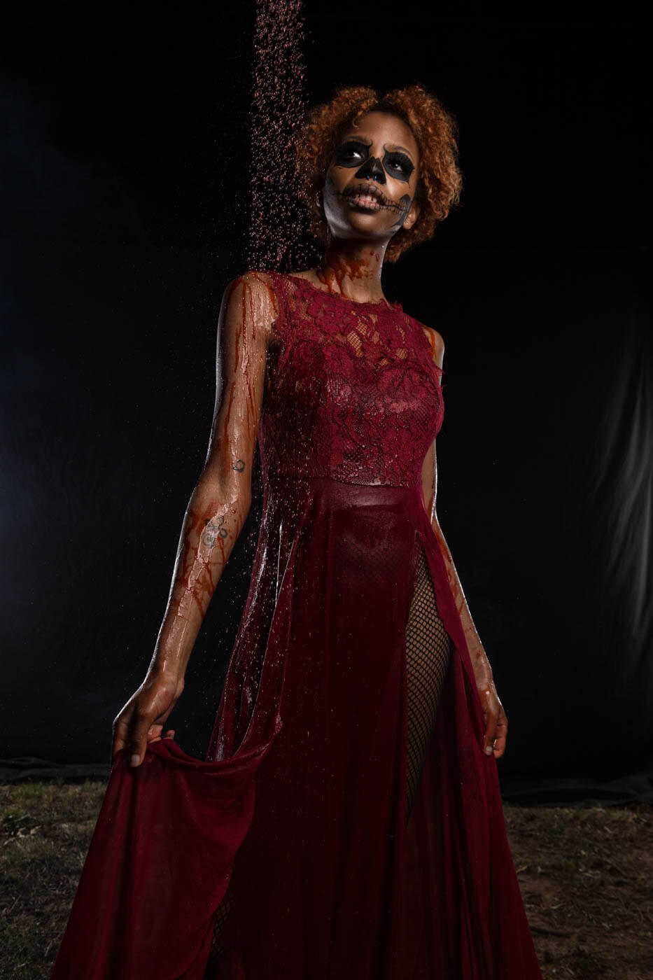 Saturn poses against a black background in a deep red lace dress with skull-inspired face paint as red liquid falls from above across her hair, shoulder, and clothing.