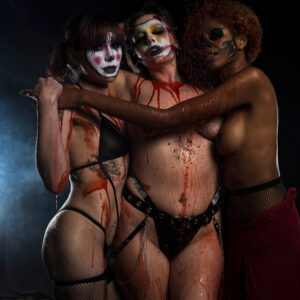Saturn, Birdie and Kody Kathleen pose together in a dark horror-themed portrait, wearing theatrical makeup and lingerie while covered in dripping red liquid beneath dramatic lighting.