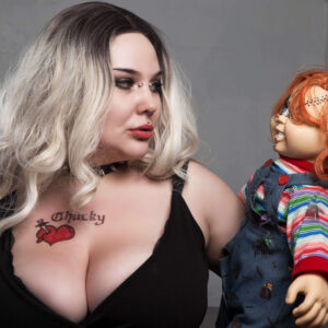 Alternative model El The Belle with platinum blonde hair, dark makeup, and a black outfit posing face-to-face with a scarred Chucky horror doll during a creative photoshoot.